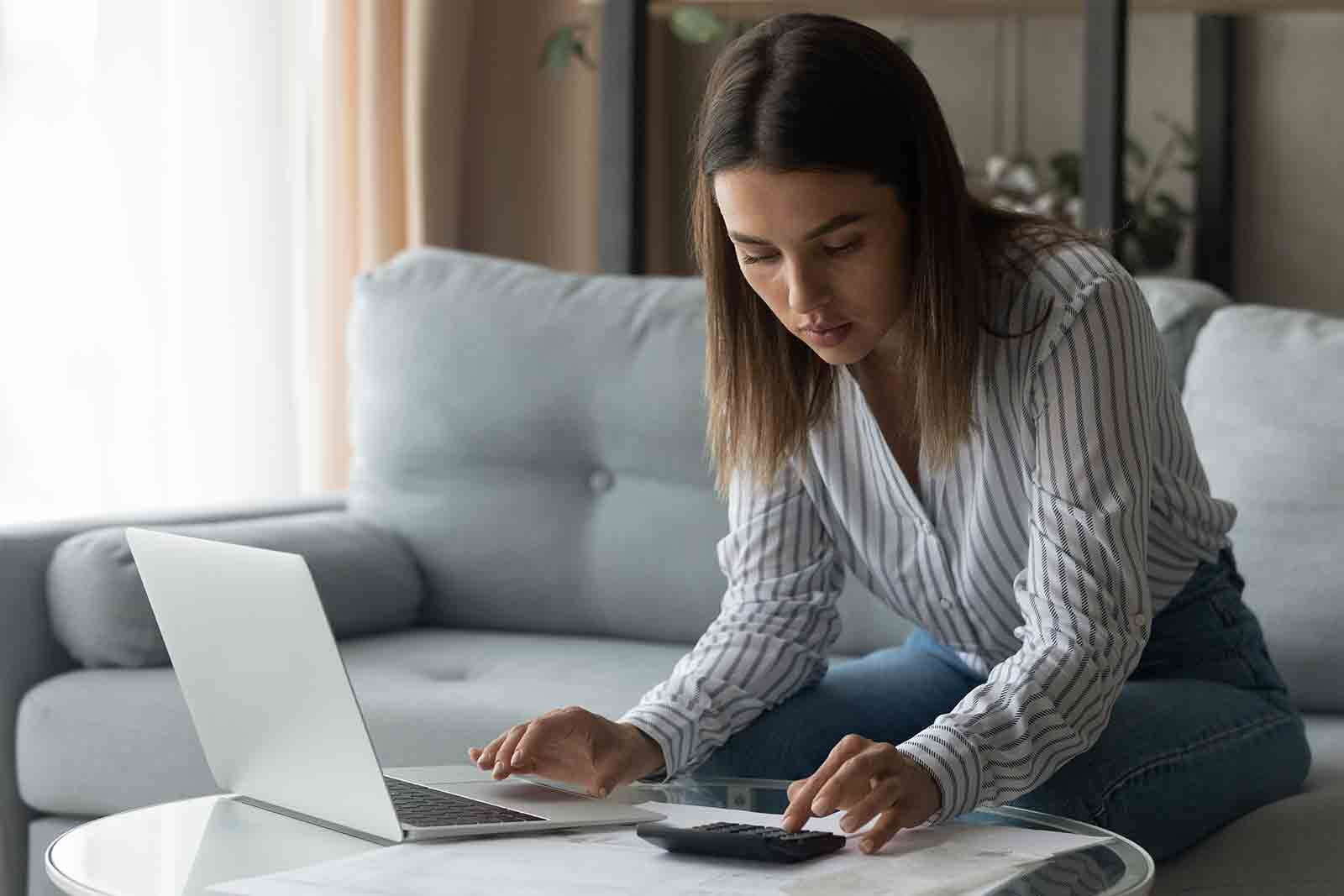 Young woman sitting in her living room with her laptop and a calculator working out her finances
